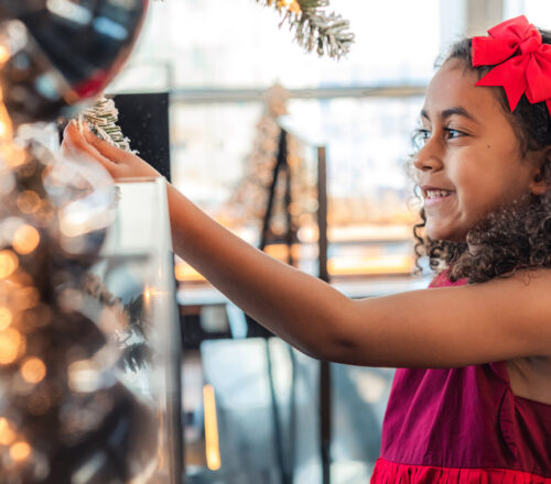 A young girl in a red dress touches the branch of a festive tree