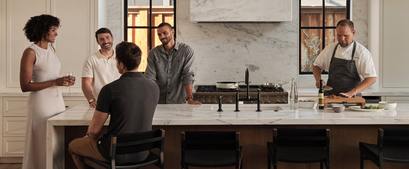 A group of people mingling in a modern kitchen while a chef prepares a meal next to them.
