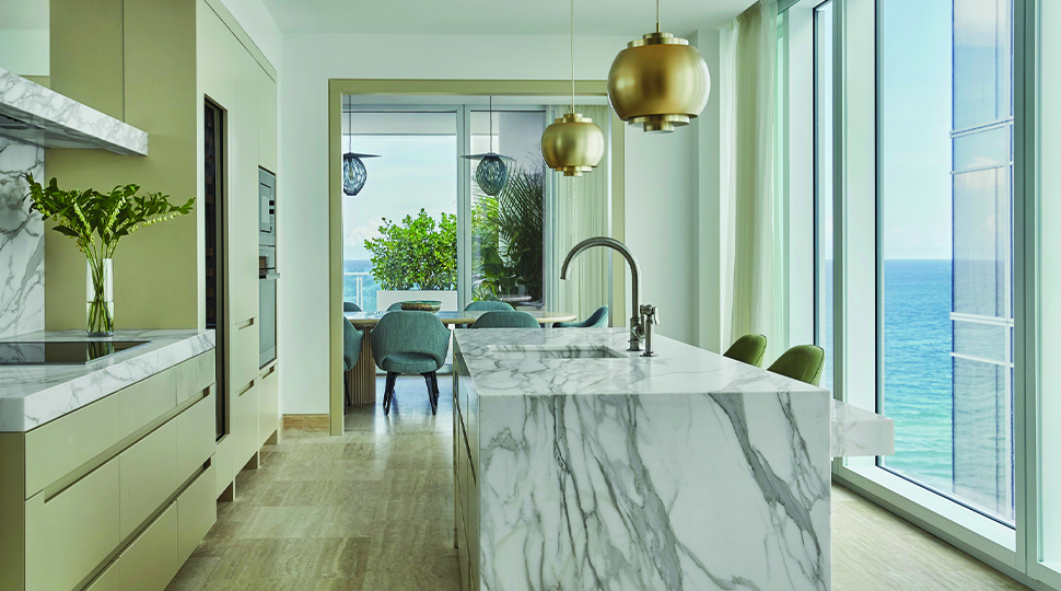 Kitchen island wrapped in white-and-grey marble with beige cabinetry on one side and floor-to-ceiling windows overlooking the ocean on the other