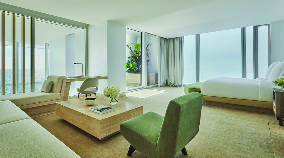 Bedroom with white bed; large seating area with a beige couch, green chair and square light-wood coffee table; floor-to-ceiling windows on one wall; and sliding glass doors that open onto a terrace