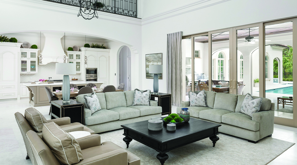 Living room with grey sofas, beige arms chairs and black coffee table with a kitchen in the background and floor-to-ceiling sliding glass doors that open onto a pool deck