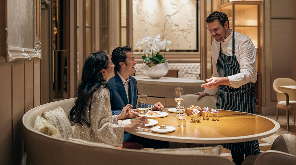 A couple sits on a curved white banquette at a round wooden table while talking to a chef standing by the table