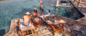 Multi-generational family of six hang out by the pool, with three adults sitting on the side of the pool on two lounge chairs and three others playing with a beach ball in the water
