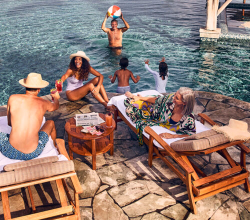 Multi-generational family of six hang out by the pool, with three adults sitting on the side of the pool on two lounge chairs and three others playing with a beach ball in the water