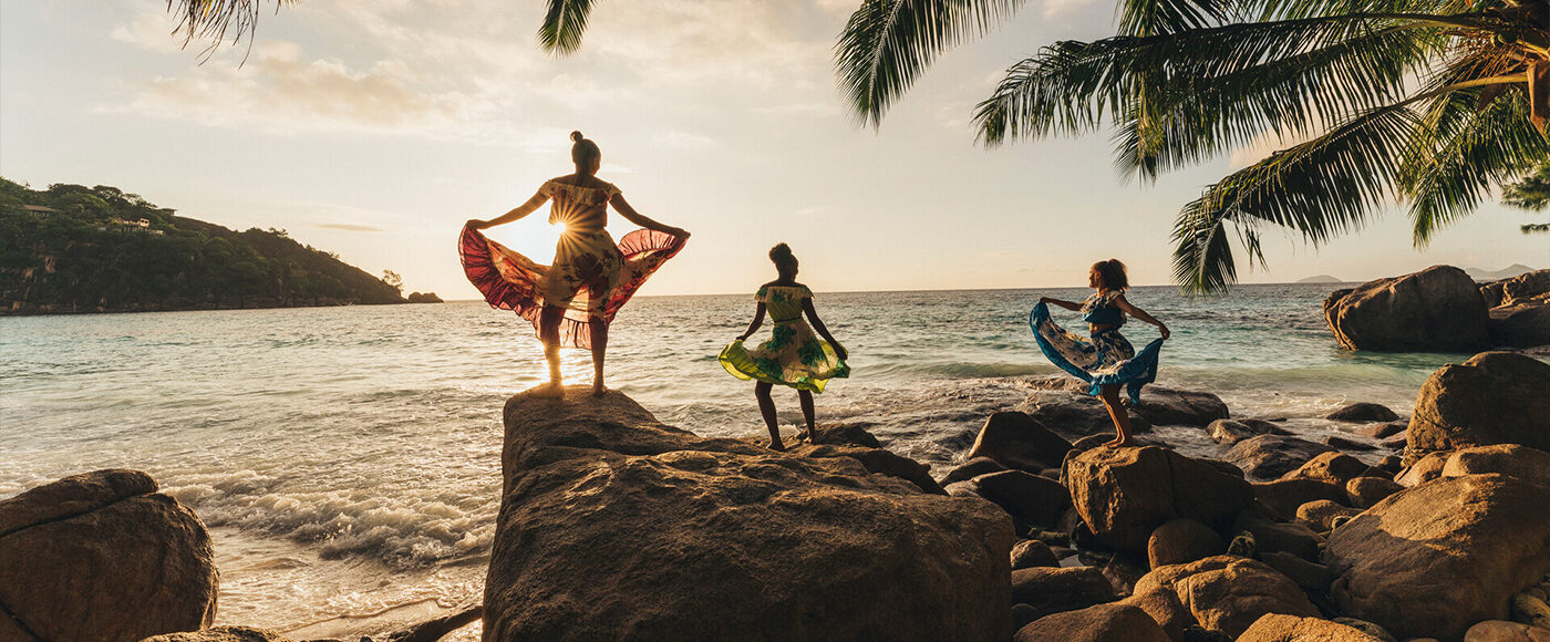 Three people stand on large boulders on the beach holding up large flowing skirts as they dance, all silhouetted by the sun