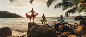 Three people stand on large boulders on the beach holding up large flowing skirts as they dance, all silhouetted by the sun