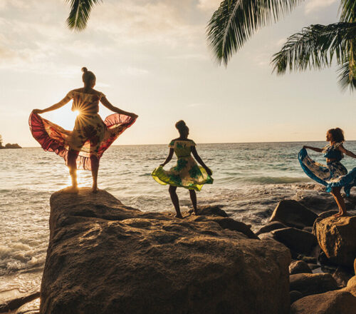 Three people stand on large boulders on the beach holding up large flowing skirts as they dance, all silhouetted by the sun