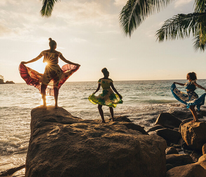 Three people stand on large boulders on the beach holding up large flowing skirts as they dance, all silhouetted by the sun
