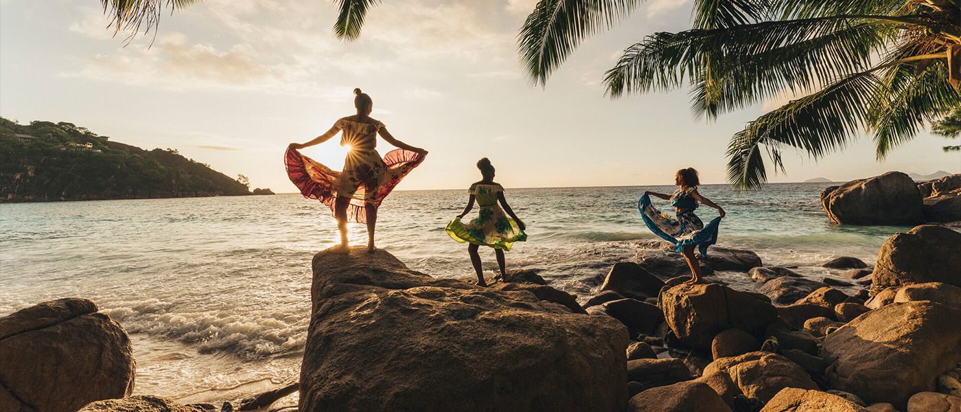 Three people stand on large boulders on the beach holding up large flowing skirts as they dance, all silhouetted by the sun