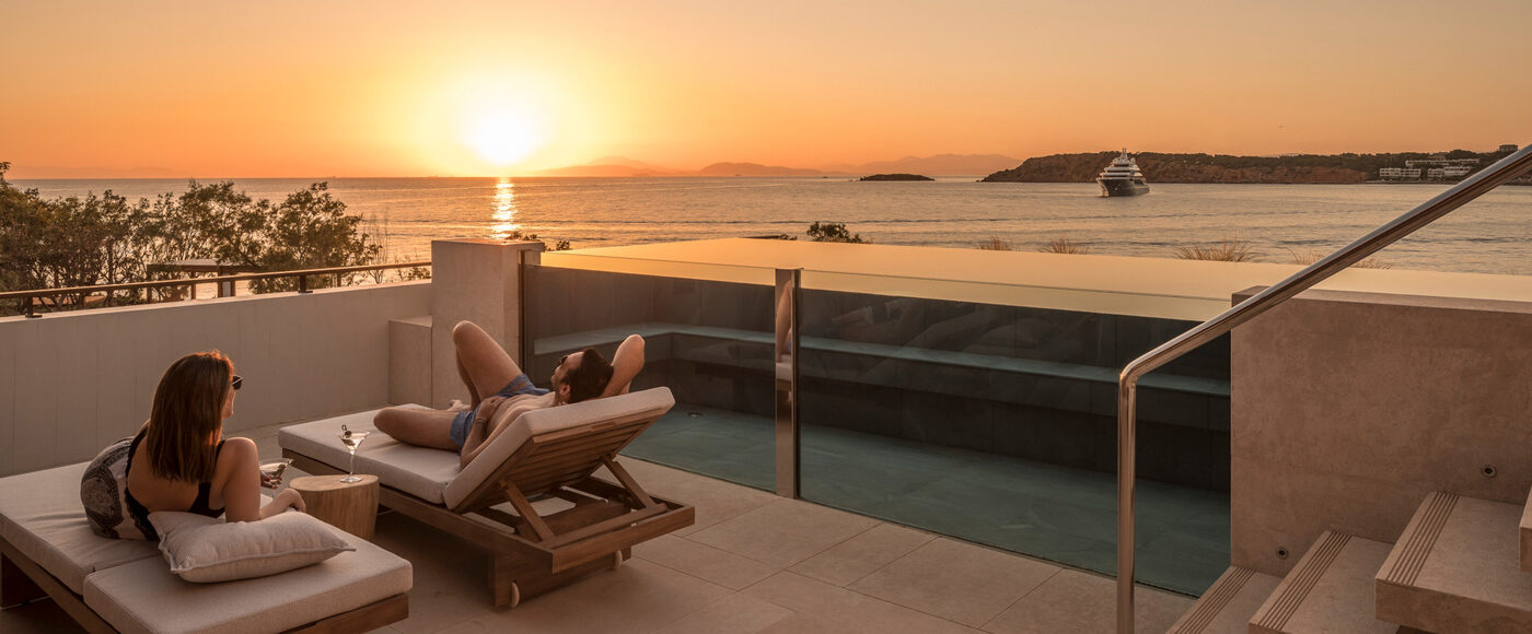 Two people recline on lounge chairs on an outdoor patio as they watch the sun set over the ocean
