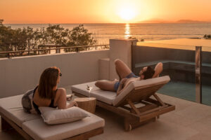 Two people recline on lounge chairs on an outdoor patio as they watch the sun set over the ocean