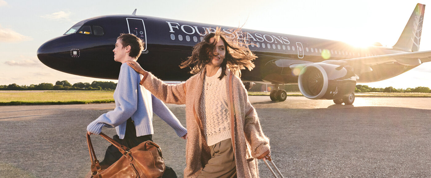 Two people wearing light-coloured sweaters and pulling rolling suitcases walk on the tarmac with a black Four Seasons branded private plane behind them