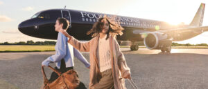 Two people wearing light-coloured sweaters and pulling rolling suitcases walk on the tarmac with a black Four Seasons branded private plane behind them
