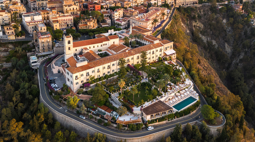Aerial view of a hotel built on top of a cliff with a city behind it and tree-covered terrain surrounding it