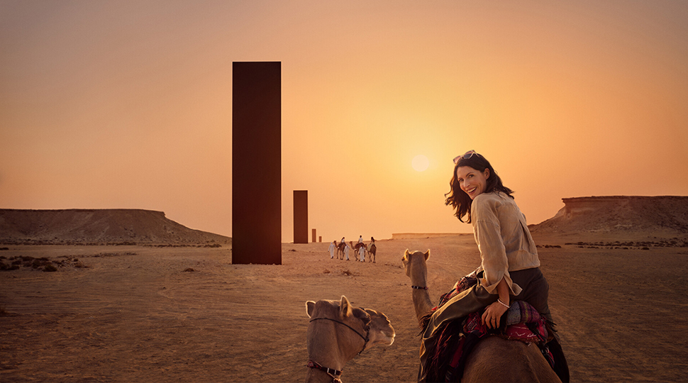 Person rising camel in the desert next to a row of black monoliths looks back over her shoulder and smiles