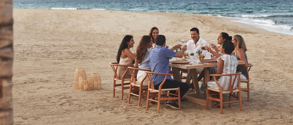 A group dines on the beach in wooden mid century modern dining chairs pulled up around a rectangular wooden dining table