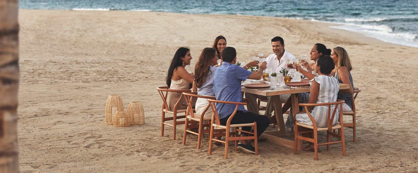 A group dines on the beach in wooden mid century modern dining chairs pulled up around a rectangular wooden dining table