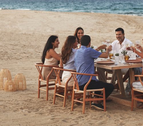 A group dines on the beach in wooden mid century modern dining chairs pulled up around a rectangular wooden dining table