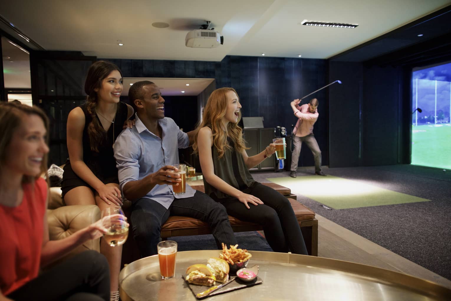 A man and three woman sip beer and cheer on a sports team in the foreground, while another man golfs in the background of a function room at four seasons hotel houston