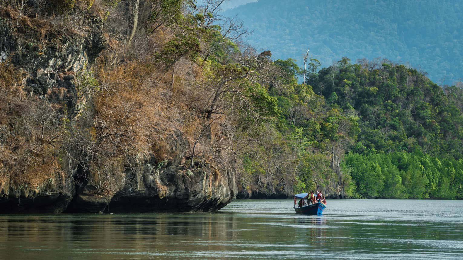 Small blue boat on river under large cliffs, trees