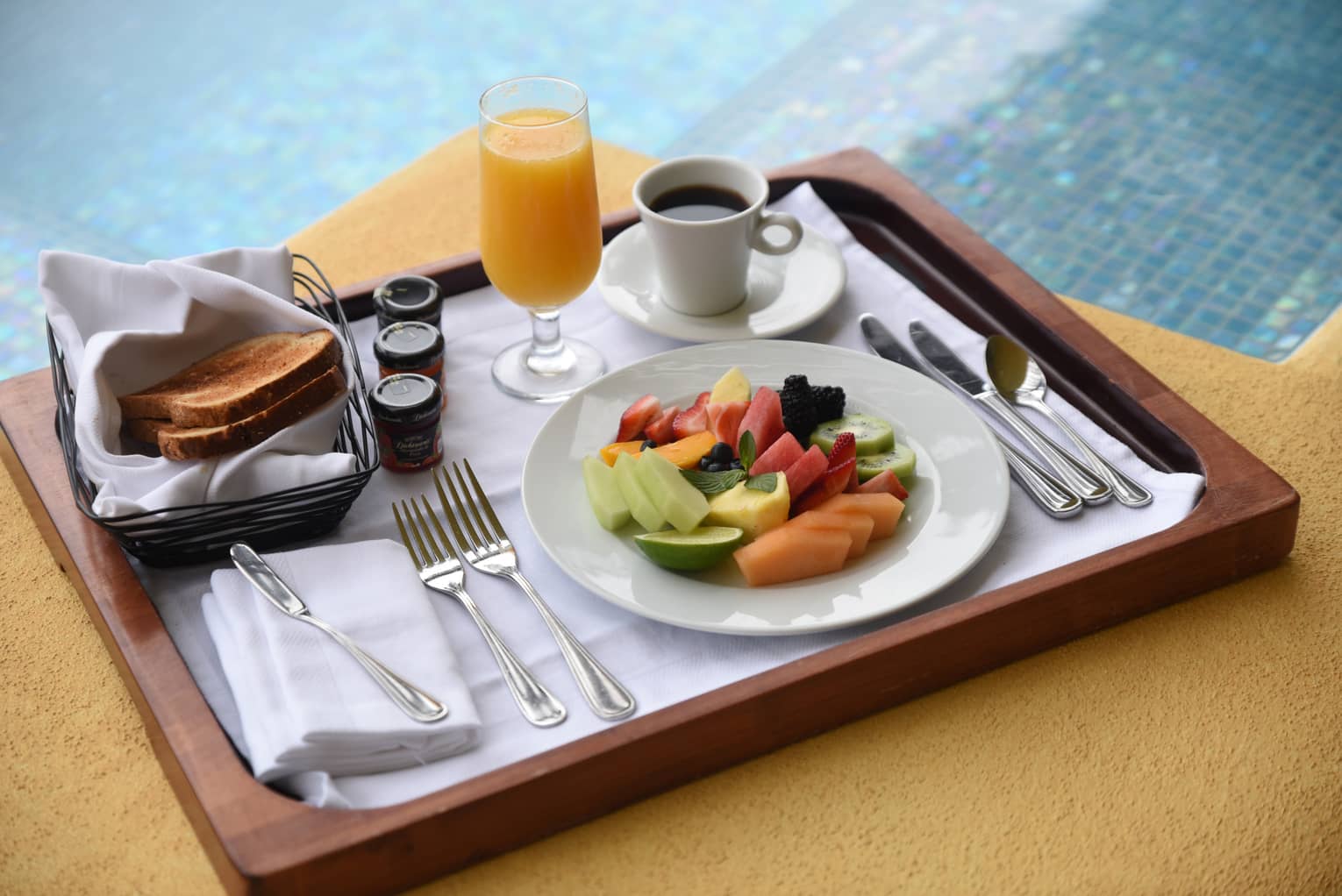 Fresh fruit slices on plate, coffee, orange juice, toast on wood tray by pool