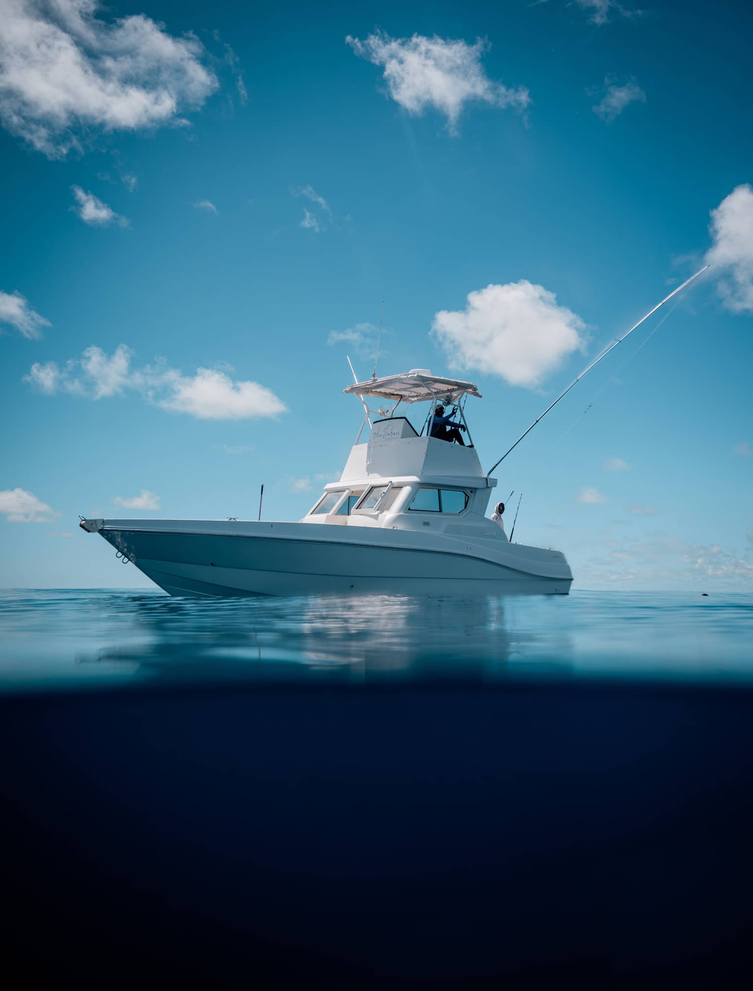 Under clear blue skies, two people aboard a centre console fishing boat; the boat's image reflected in the still water below.