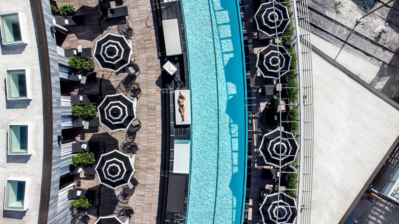 An aerial view looking down on a long, slightly curved pool and surrounding deck area, which includes tables with umbrellas, potted greenery and places where guests can recline next to the pool