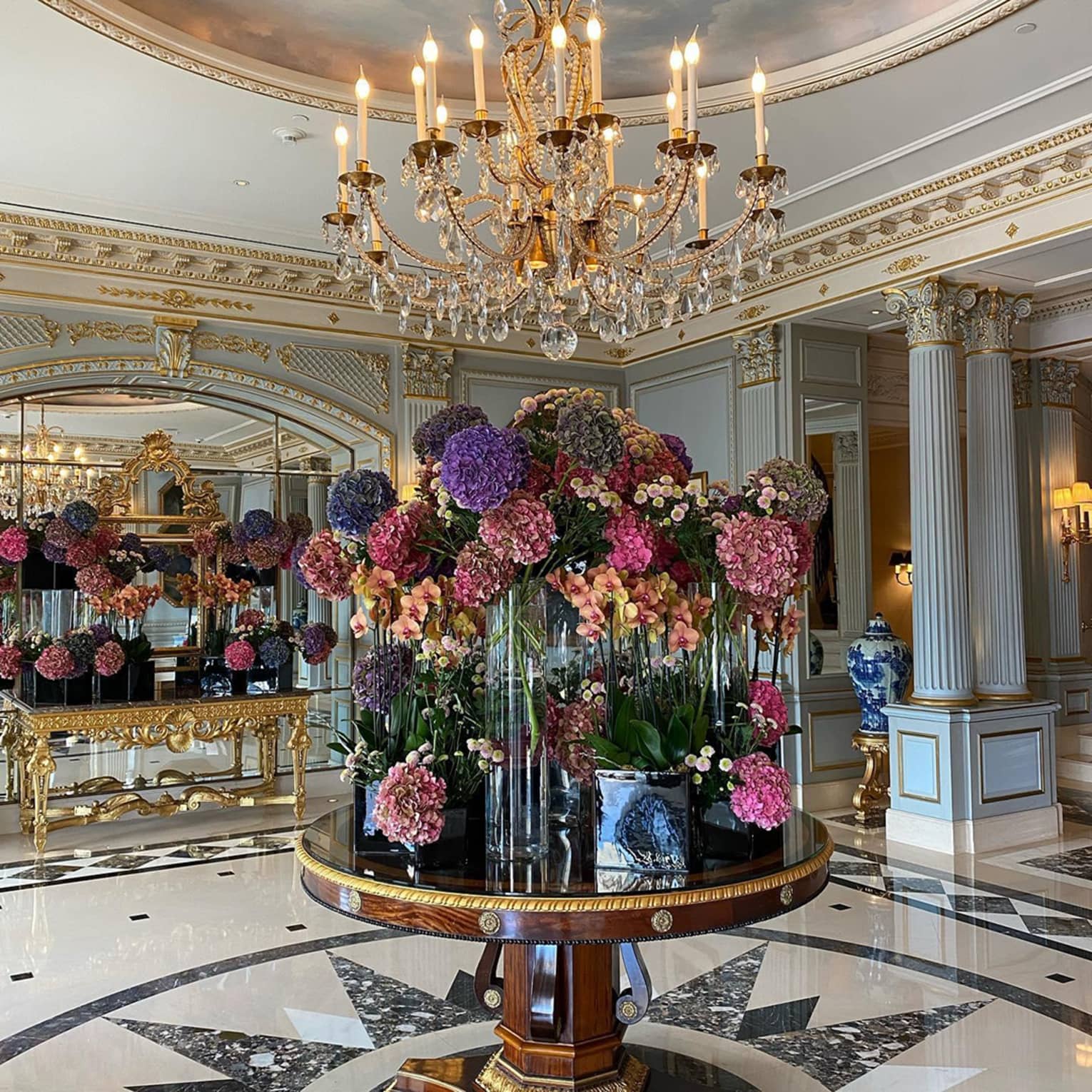 Five-star luxury hotel lobby with a chandelier above a round table adorned with pink, purple and peach floral arrangements, mirrored walls and gold accents