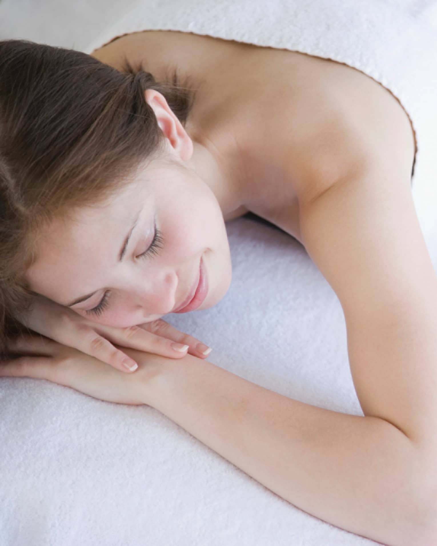 Smiling woman rests on spa table with head on arms beside ceramic bowl with water, flower petals