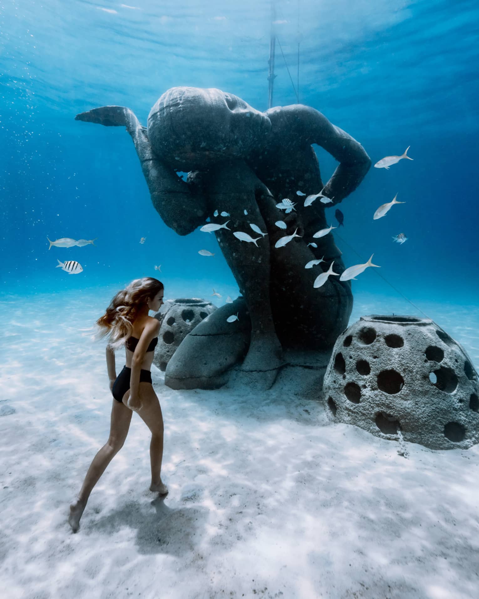 Person walking underwater near a large statue surrounded by fish, in a clear, blue ocean setting