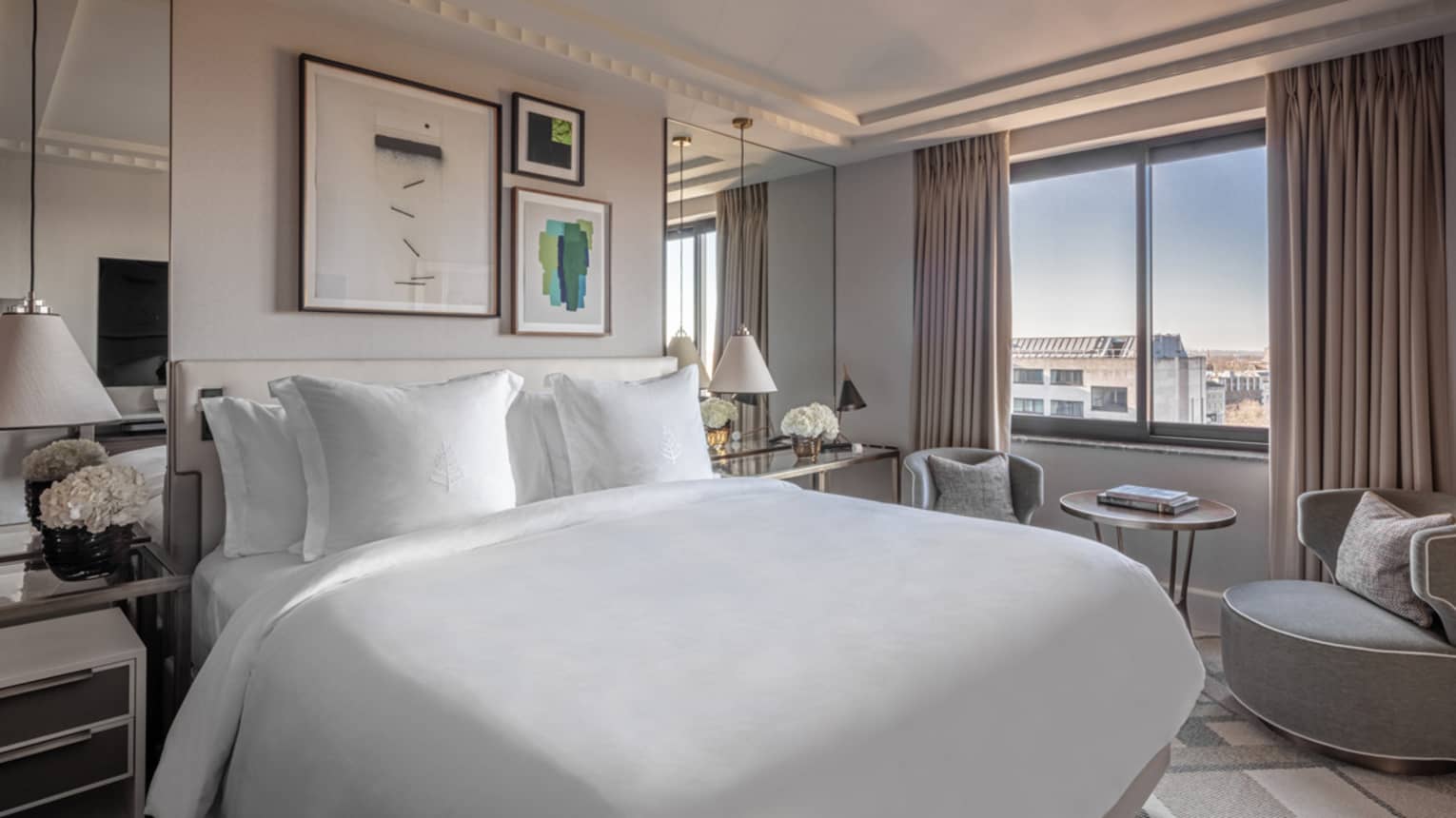,Superior Room at Four Seasons London Park Lane in Mayfair, offering a blend of comfort and luxury. The room features a large, king-sized bed covered in crisp white linens, surrounded by tasteful decor, including muted gray curtains, elegant lamps and a selection of framed artworks on the walls. A comfortable seating area by the window provides a relaxing space with a curved armchair and a small table, perfect for enjoying the city views. The room's modern and understated elegance is emphasized by its clean lines and soft colour palette.