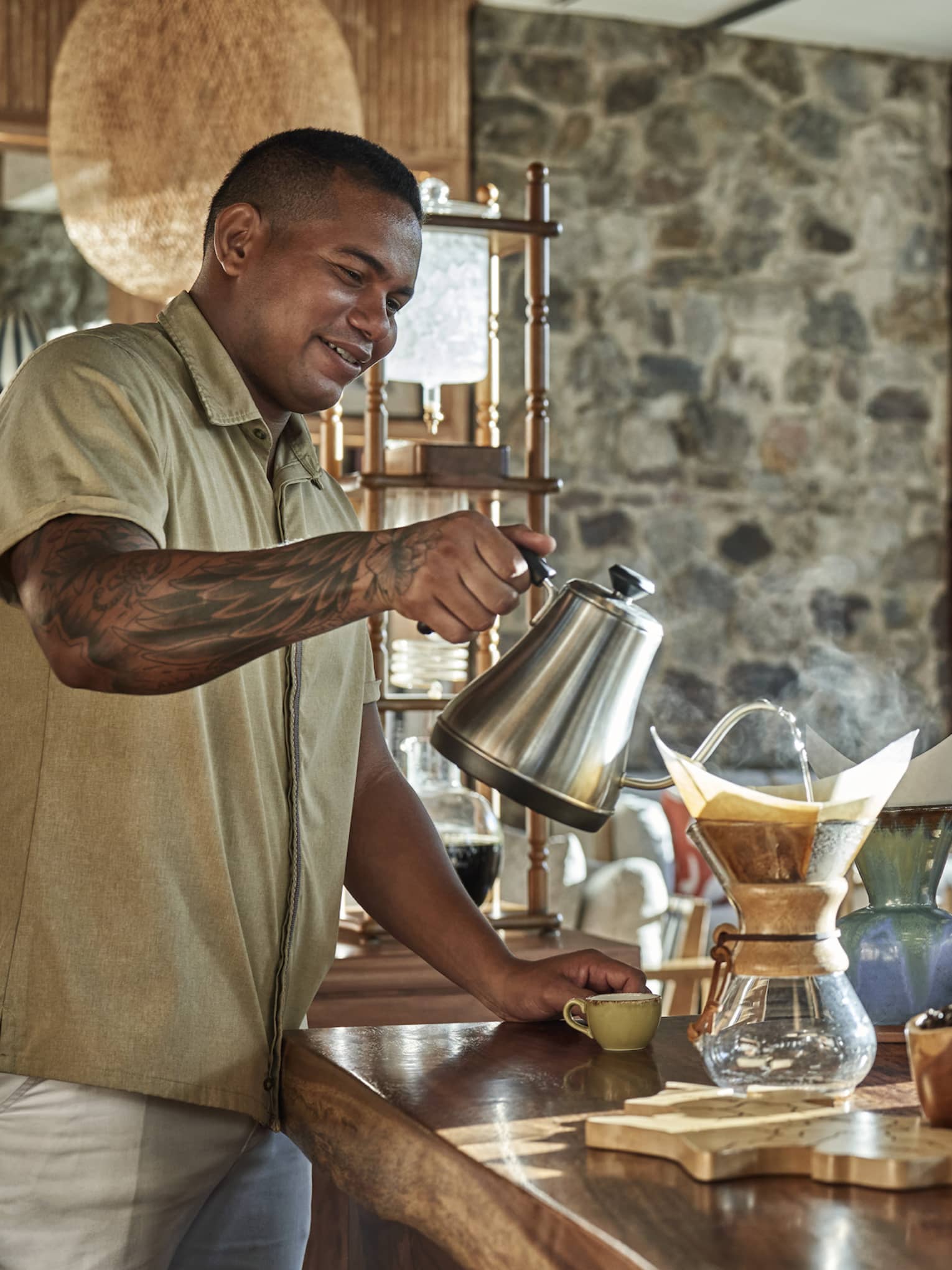 A man sits next to a wooden counter smelling a fresh cup of coffee, a woman stands next to him and another man stand on the opposite side pouring water into a chemex