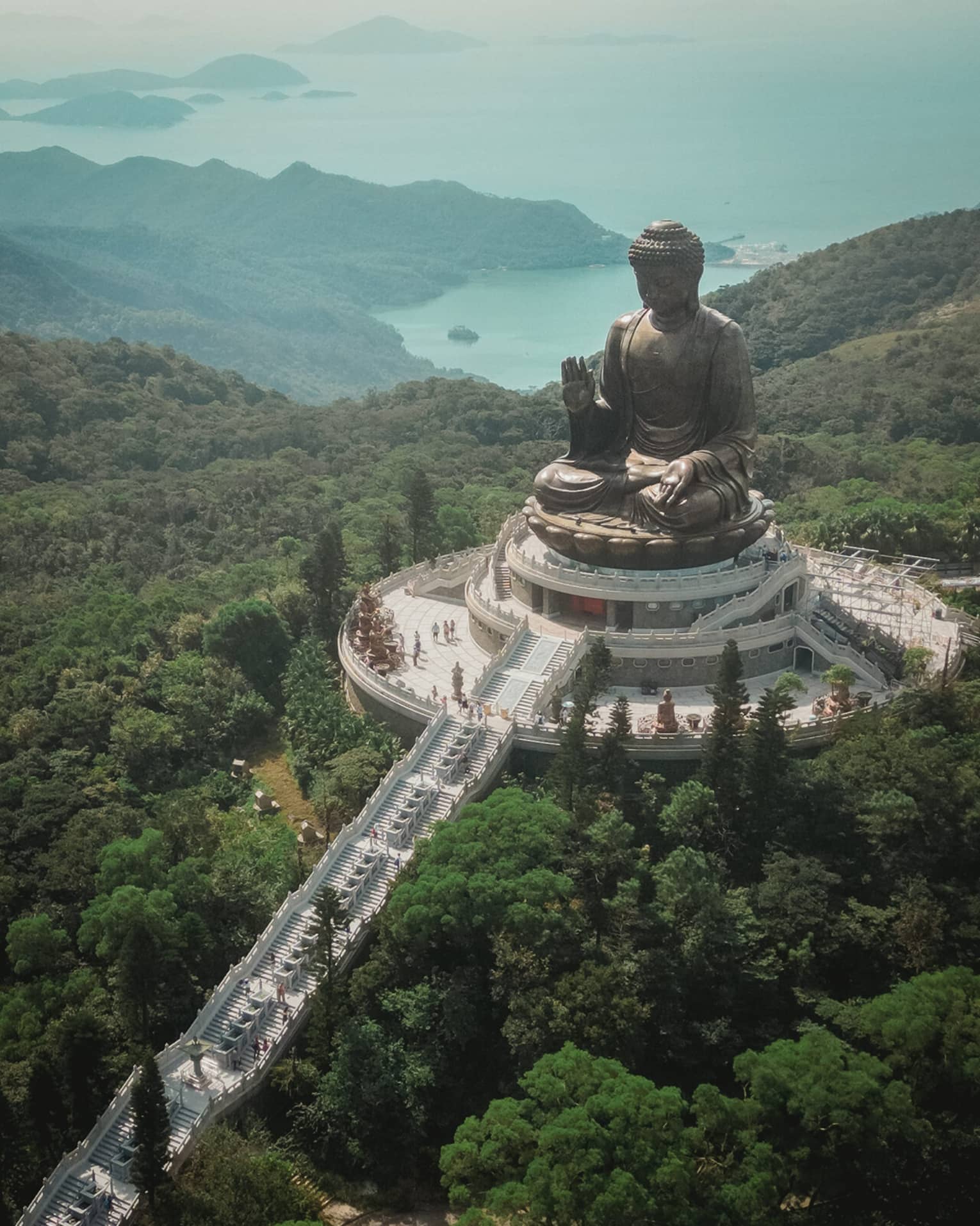 Aerial view of a giant bronze Buddha statue at Po Lin Monastery, surrounded by greenery and mountains. A long staircase leads to the statue on a platform