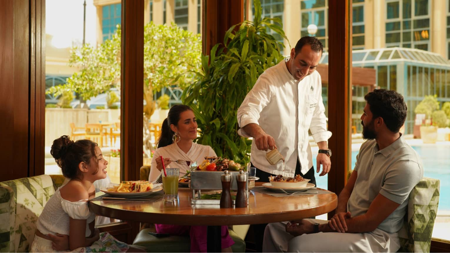Family of three seated at round wooden table at poolside restaurant are served by chef in white coat