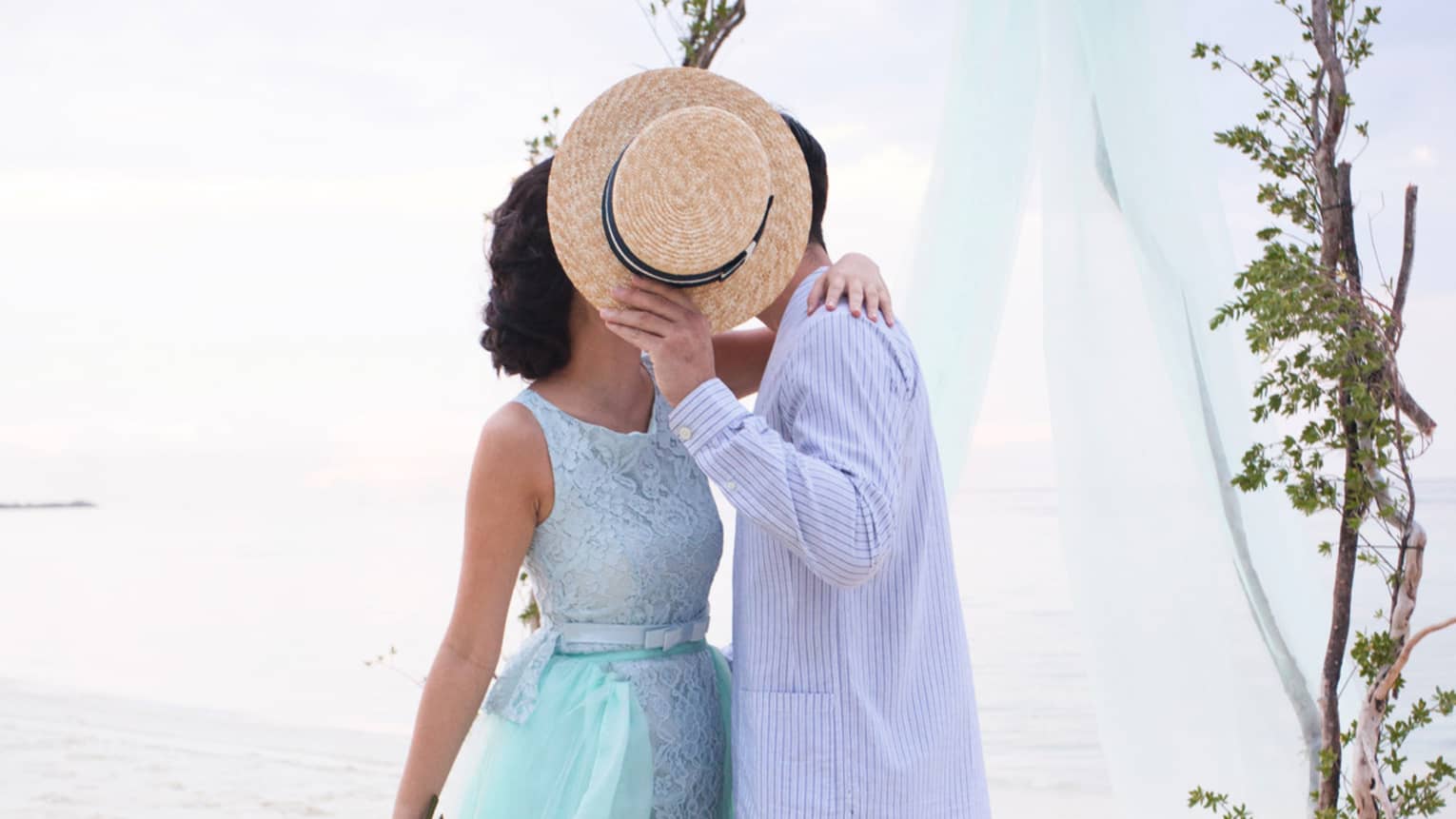 Woman and man kiss behind straw hat at outdoor wedding ceremony