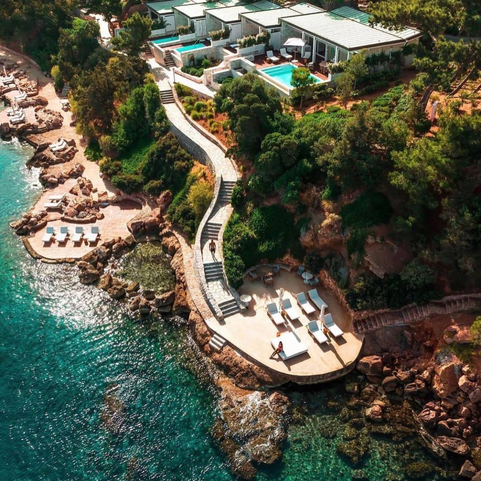 Aerial view of Four Seasons Astir Palace Hotel Athens, with modern villas, private swimming pools, and a long staircase leading to a patio with lounge chairs, next to clear, blue waters
