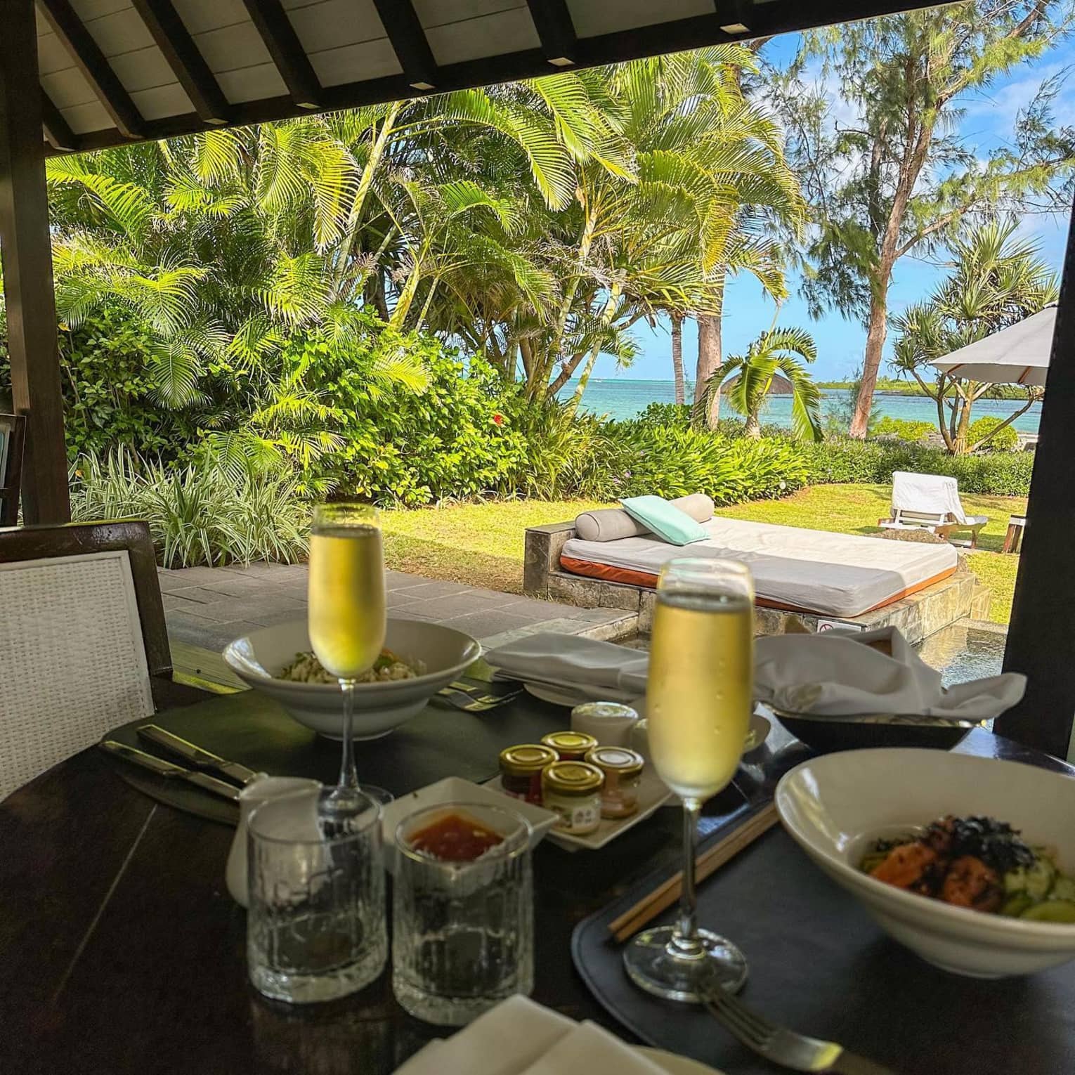 An outdoor table with food and two chilled glasses of champagne overlooking a pool, daybed and ocean view