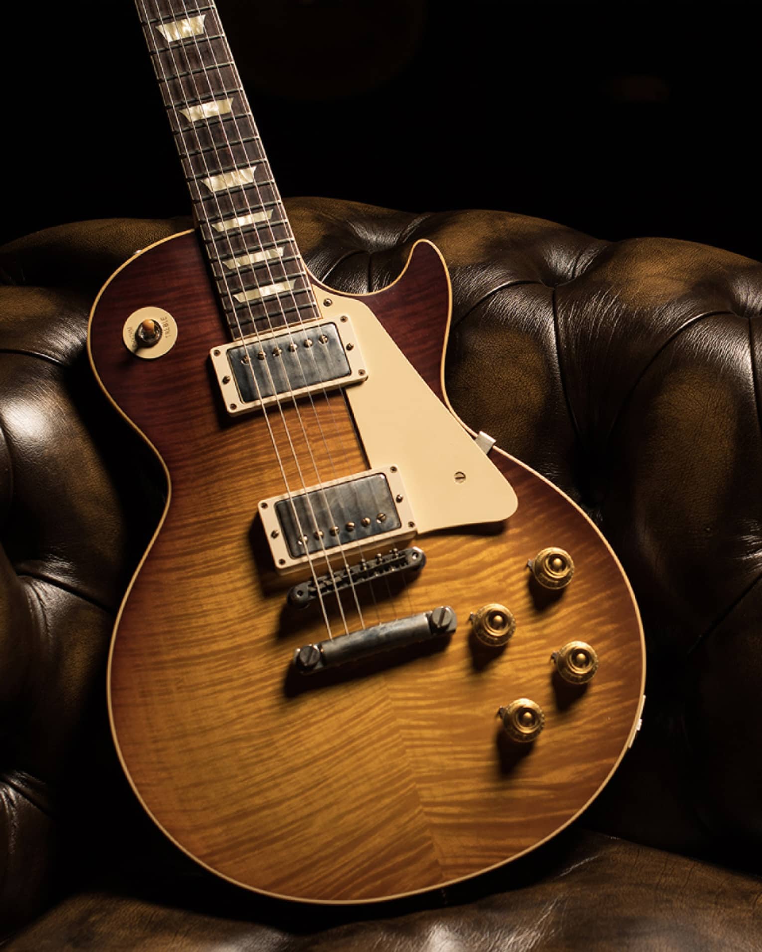 Close-up of a sunburst-finished electric guitar, its glistening body leaning diagonally against a brown leather tufted couch.