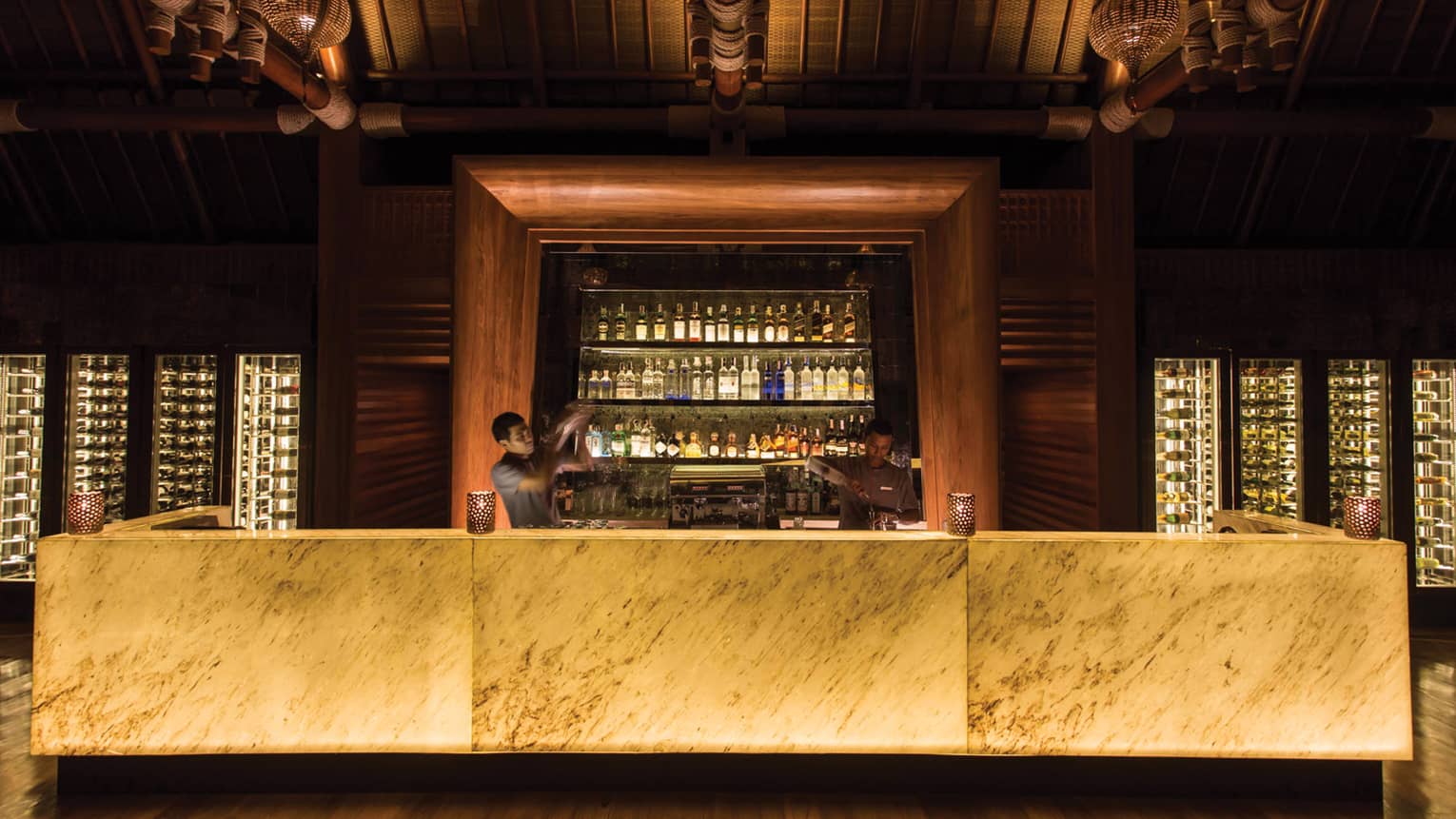 Two bartenders mix cocktails behind the dimly-lit bar, liquor bottles on display behind them