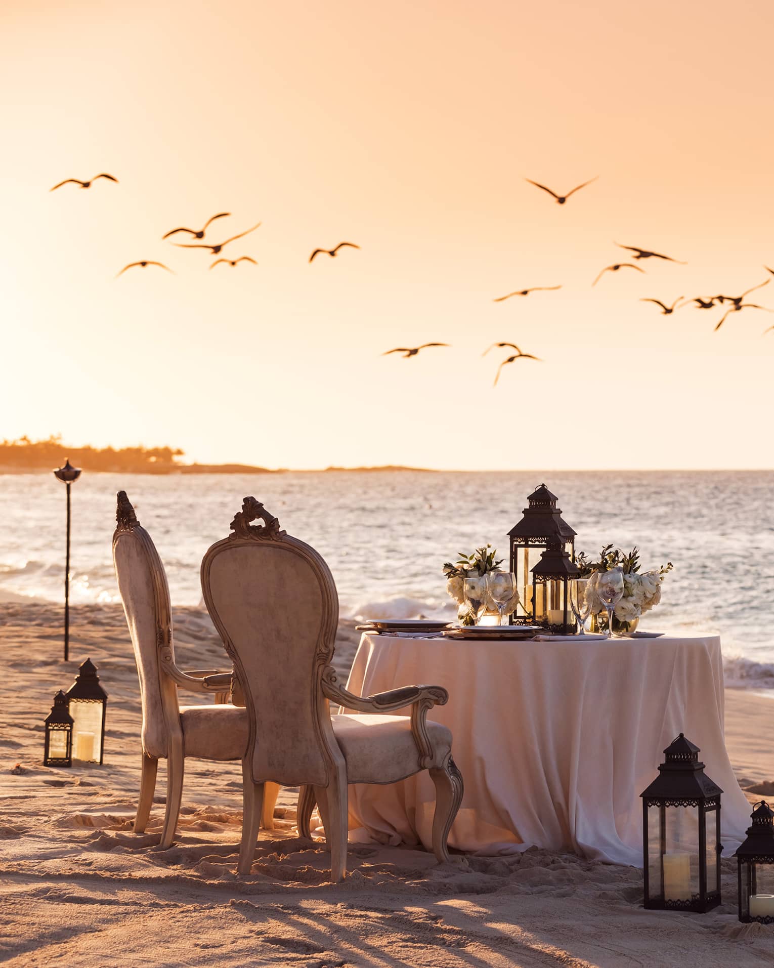 Romantic beachside dining setup with vintage chairs, a table decorated with flowers and lanterns and birds flying overhead