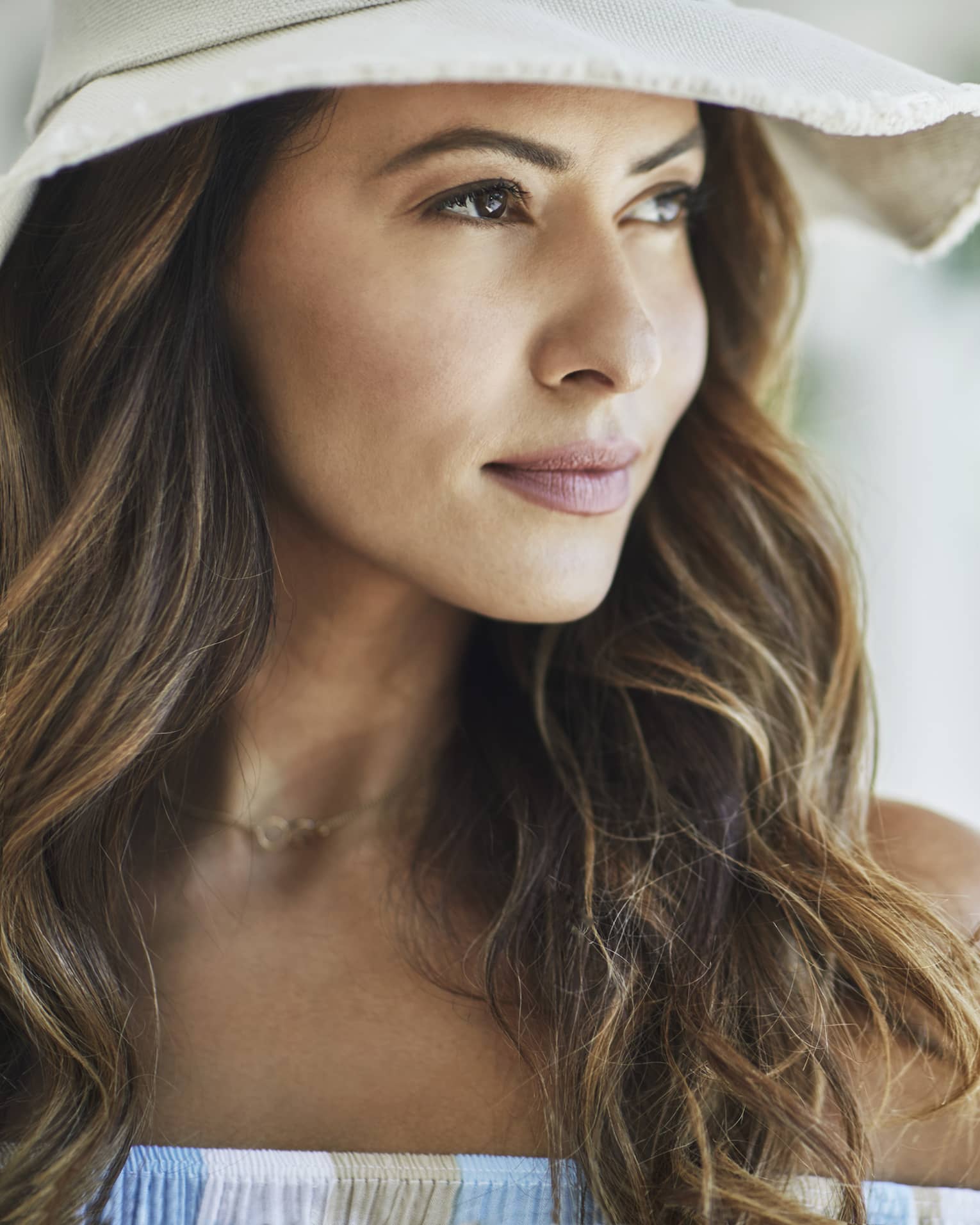 A woman with wavy brown hair and a white floppy hat looks off into the distance