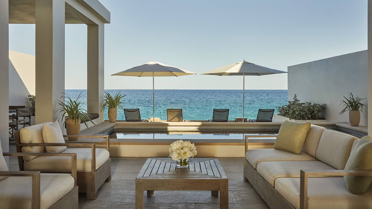 Outdoor terrace with cushioned seating, a wooden table, two umbrellas and ocean views beyond a private pool
