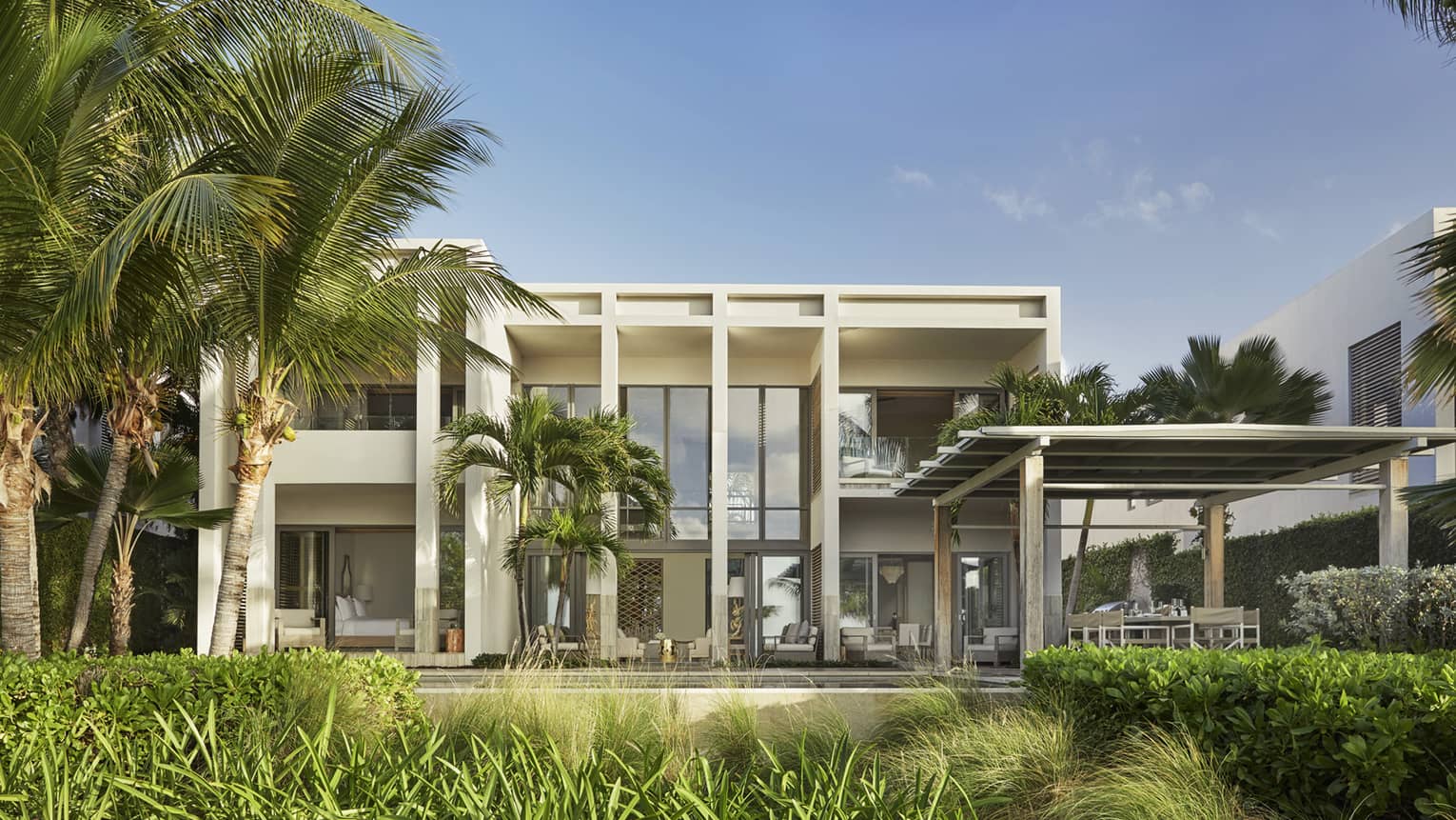 Modern beachfront villa with large glass windows, palm trees and a shaded outdoor seating area.