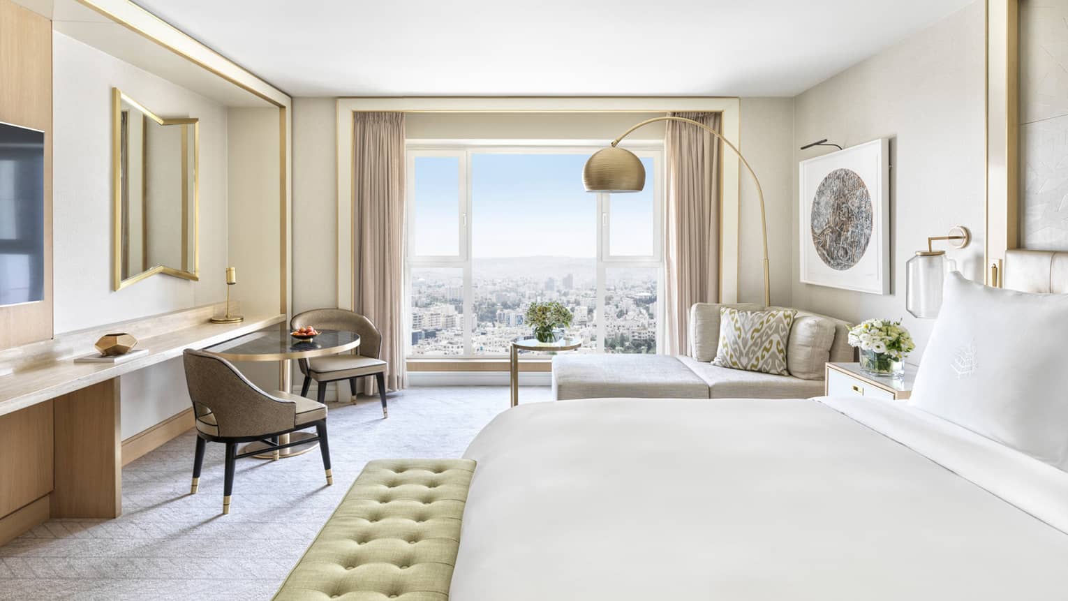 Hotel room with white king bed, light green cushioned bench, two taupe chairs, chaise, wall of windows