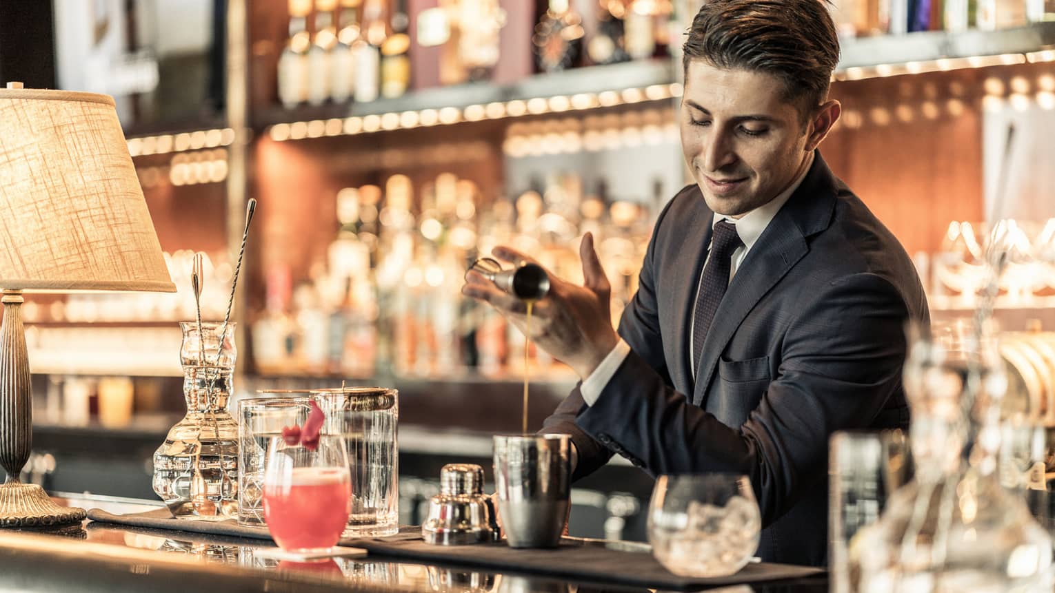 Bartender behind bar, pouring liquid from jigger into metal shaker among bar accessories