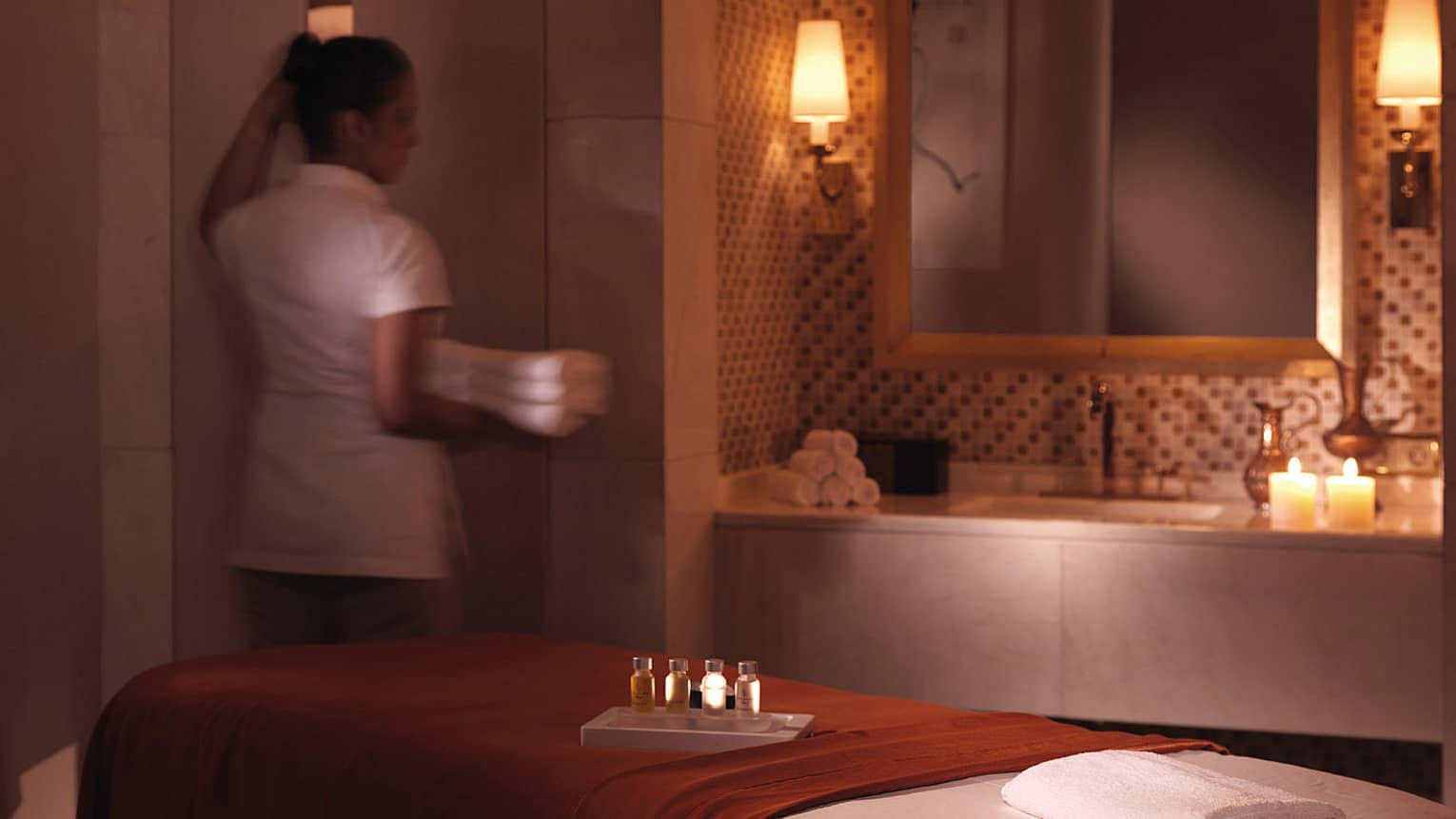 Spa attendant in white uniform holds three folded white towels by massage table with blanket, tray with small oils