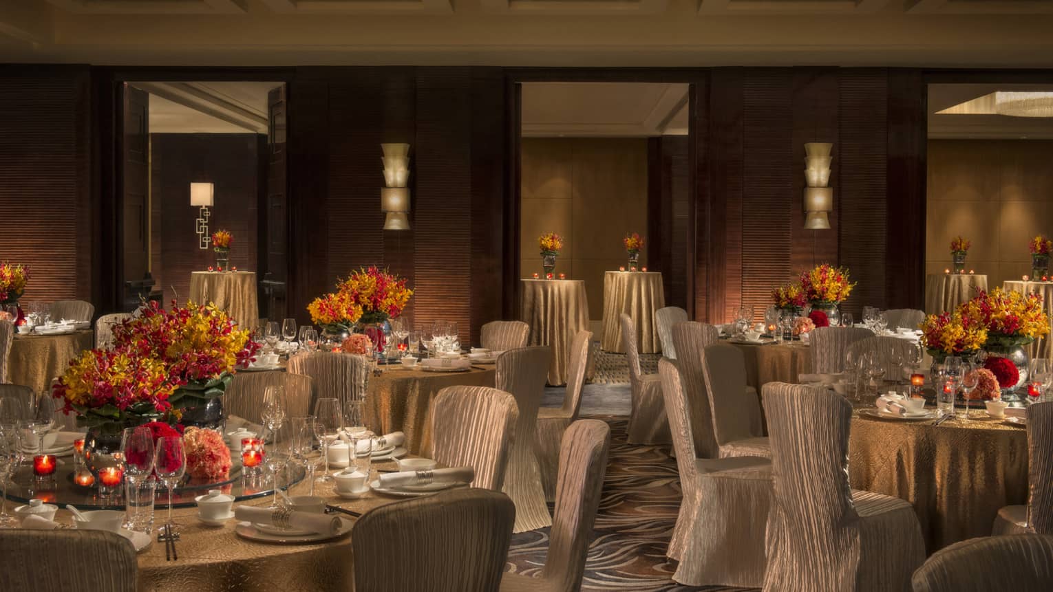 Dimly-lit ballroom with silk covered chairs around reception tables with orange flowers, candles