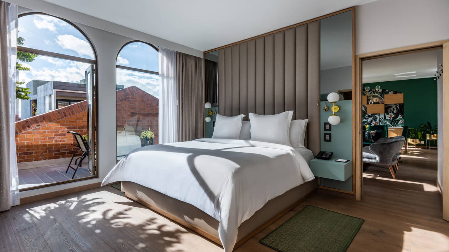 Guest room with bed and white linens, floor-to-ceiling windows overlooking the terrace, living/dining area just visible around the partition