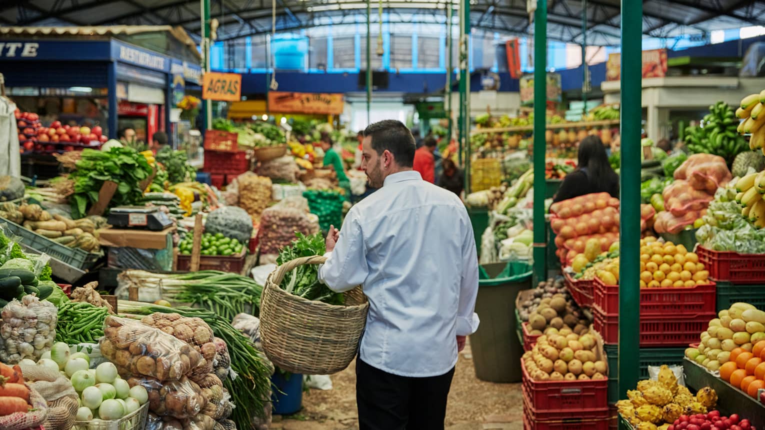 Back of man with beard carrying wicker basket through fruit market aisle, produce stacked on shelves