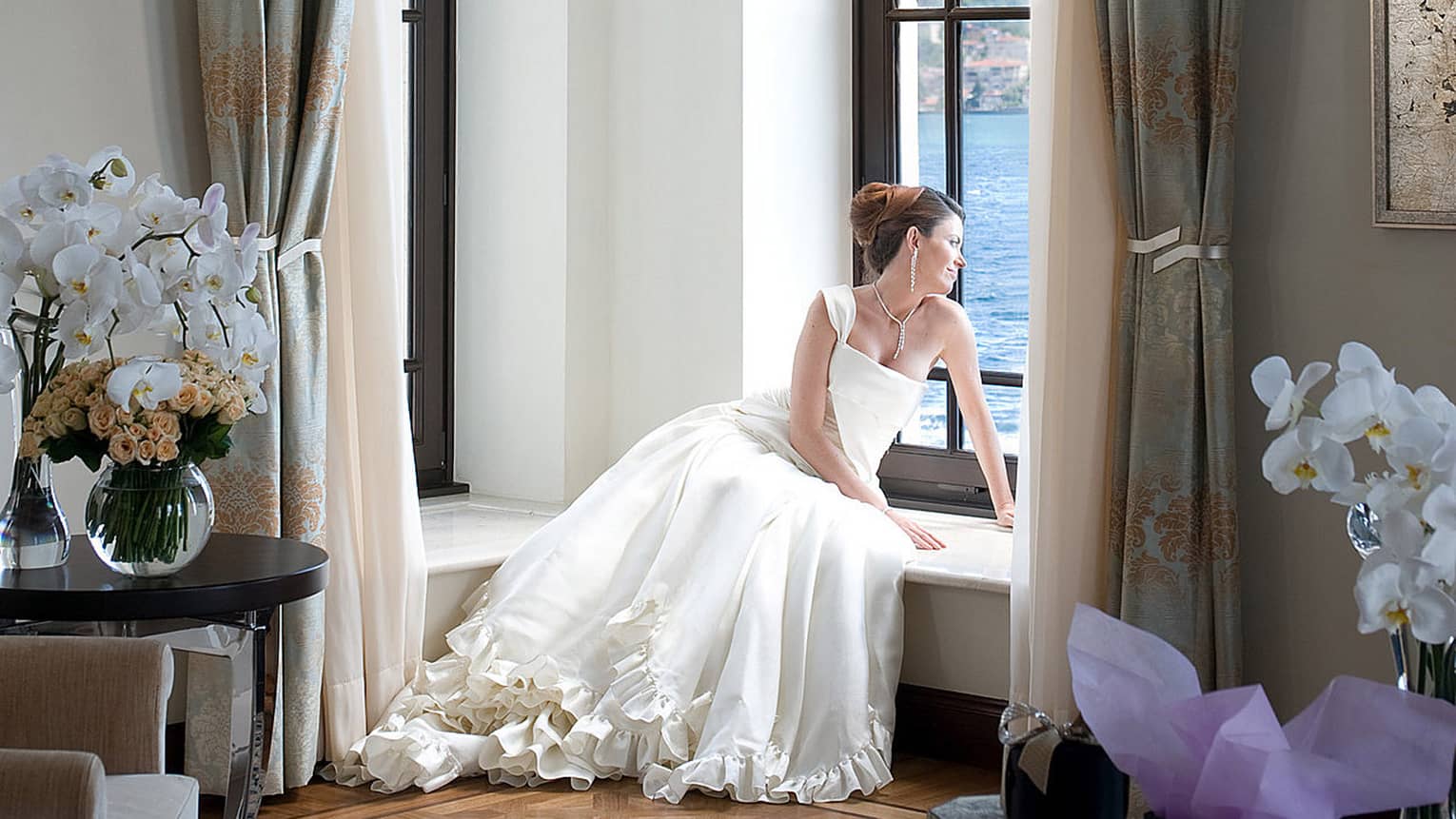 Bride wearing elegant white wedding dress lounges in window sill in room with white orchids